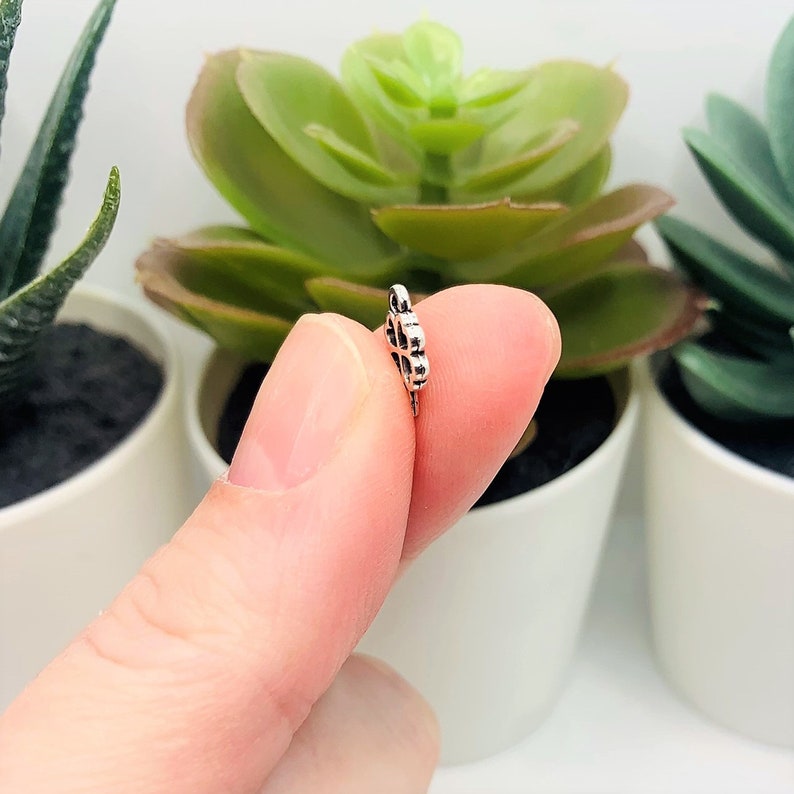 May include: A silver charm in the shape of a flower with a detailed design. The charm is being held by a finger.