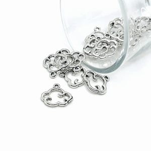 May include: A group of silver metal cloud charms with a decorative design. The charms are in a glass bowl and some are spilling out.
