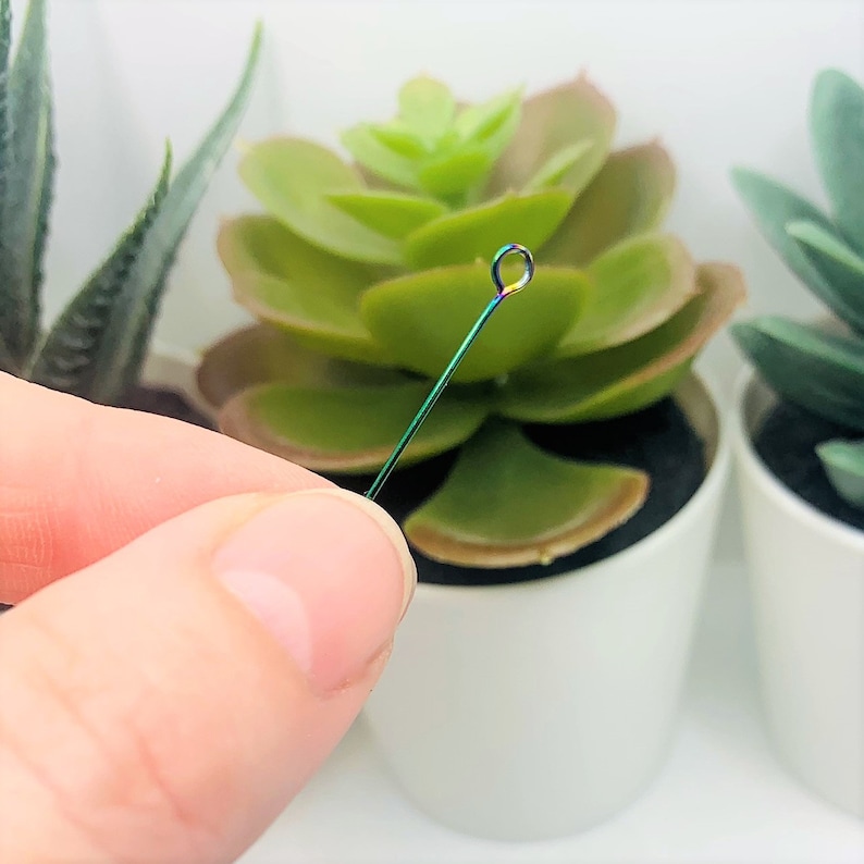 May include: A thin, iridescent, green wire with a small loop at the top. The wire is held in a hand against a blurred background of potted plants.