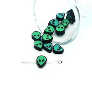 May include: A collection of black and green alien-shaped beads. The beads have a green face with two eye holes and a black outline. One bead is strung on a silver wire. The beads are scattered near a clear glass container.