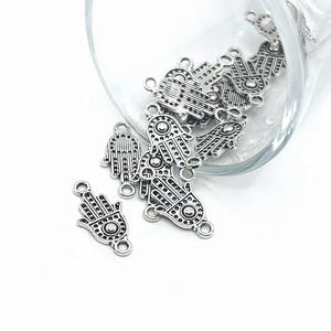 May include: A close-up of a glass bowl filled with silver-toned metal charms shaped like the Hamsa hand, a symbol of protection and good luck. The charms have intricate details and are perfect for jewellery making or crafting.
