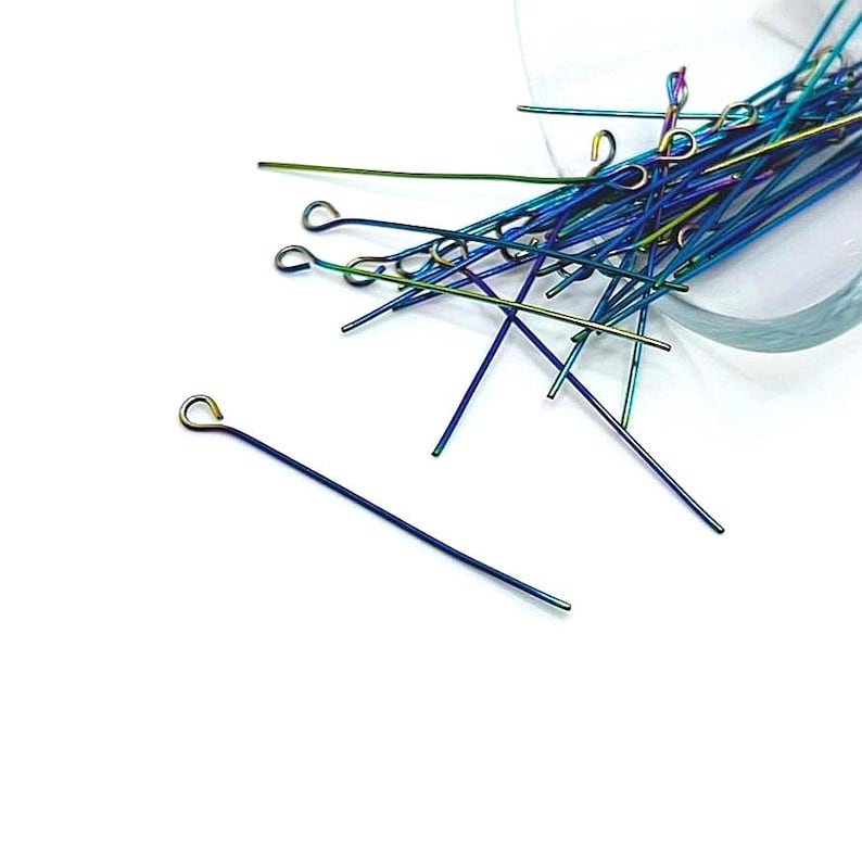 May include: A pile of iridescent blue, green, and purple head pins with a loop at one end. The pins are scattered on a white surface.