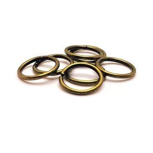 May include: A group of six gold-toned metal jump rings. The rings are all the same size and are arranged in a pile.