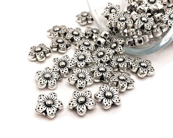 May include: Silver-colored metal flower beads scattered on a white surface. The beads are in the shape of five-petal flowers with detailed textures. The beads are likely for jewelry making or other craft projects.