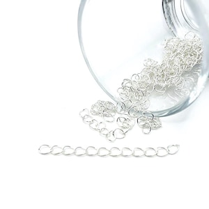 May include: Close-up of silver-colored jewelry chain links, some scattered and some in a clear glass container. The links are small and delicate, with a bright, reflective surface. The chain is displayed against a white background.
