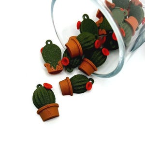 Pode incluir: Um grupo de pequenos amuletos de cacto verde com detalhes de flores vermelhas, que se espalham de um frasco de vidro. Os amuletos são feitos de plástico e têm uma aparência realista.