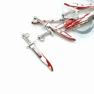 May include: A group of silver-toned, bloody knife charms with red paint accents. The charms have a loop at the top for attaching to a necklace or bracelet.