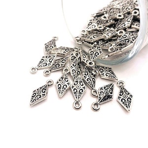 Puede incluir: Encantos de plata en forma de diamante con diseños intrincados y un bucle para colgar. Los encantos están dispersos sobre una superficie blanca.