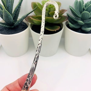 May include: Silver bookmark with an ornate design and a hook at the top. The bookmark is held in a hand.