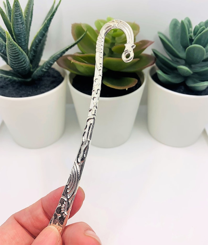 May include: Silver bookmark with an ornate design and a hook at the top. The bookmark is held in a hand.