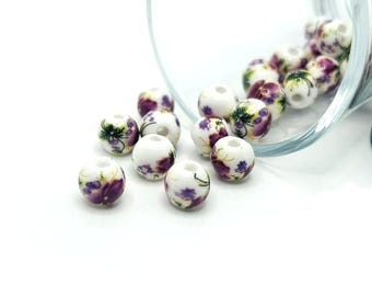 May include: White ceramic beads with purple and green floral patterns are scattered on a white surface. The beads are round with a hole through the center. Some beads are still inside a clear glass container.