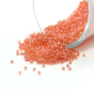 Peut inclure: Gros plan sur un récipient en verre transparent renversé, déversant une masse de petites perles de rocaille orange-rose. Les perles sont éparpillées sur une surface blanche. Idéal pour la création de bijoux et l'artisanat.