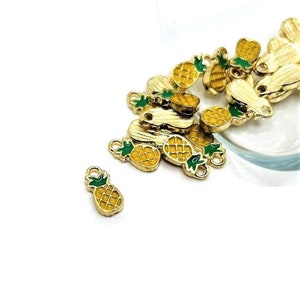 May include: A group of gold-colored pineapple charms with green tops. The charms are scattered on a white surface.