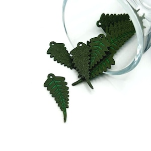 May include: Dark green fern leaf charms with a textured surface and a small hole at the top. Several charms are scattered on a white surface, with some spilling from a clear glass container. The charms have a detailed, realistic appearance.