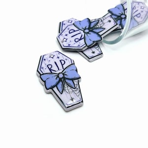 May include: Purple and white coffin-shaped charms with a blue bow and the text "RIP" on each charm. The charms have a spiderweb design and glitter accents.