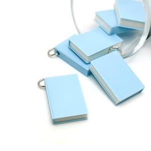 May include: Several small, pale blue book-shaped charms with silver key rings. The charms have white pages and are scattered on a white surface, with some spilling from a clear glass.