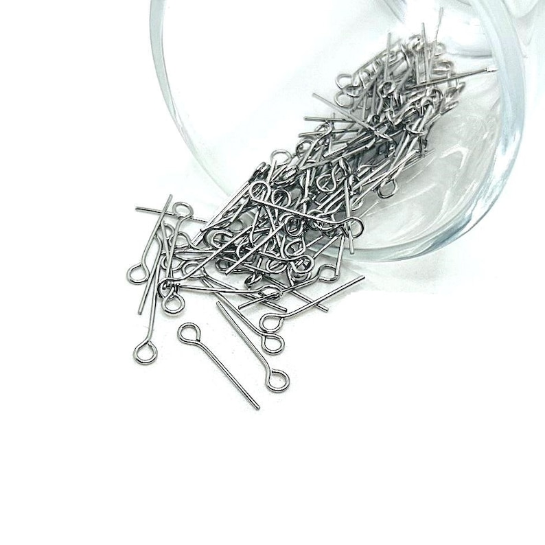 May include: A glass jar filled with silver-toned head pins, spilling out onto a white surface.