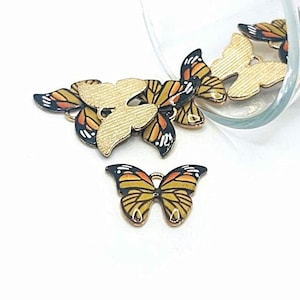 May include: A group of gold-colored metal butterfly charms with black and orange painted wings. The butterflies are scattered on a white surface.