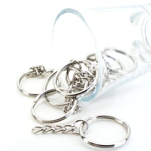 May include: A collection of shiny silver key rings and chains spilling from a clear glass container. The key rings are round and connected by small chains, creating a bright, metallic display against a white background.