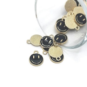 May include: A collection of gold-colored metal charms with black enamel smiley faces. The charms have a textured, circular design and a small loop for attaching to jewelry or other crafts. Some charms are inside a clear glass container.