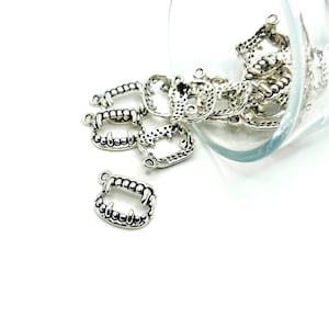 May include: A pile of silver-toned metal charms shaped like fangs, with a textured surface and a small loop at the top. The charms are spilling out of a clear glass jar.