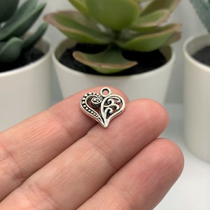 May include: Silver charm in the shape of a heart with intricate filigree details. The heart is ornate and has a loop at the top for attaching to a necklace or bracelet.