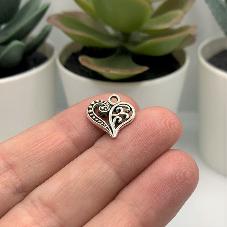 May include: Silver charm in the shape of a heart with intricate filigree details. The heart is ornate and has a loop at the top for attaching to a necklace or bracelet.