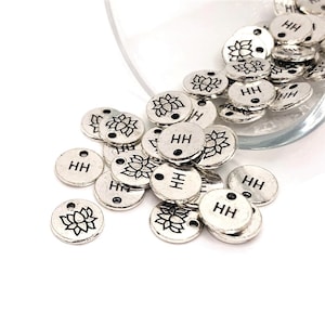 May include: A pile of silver metal charms with a lotus flower design and the letters "HH" engraved on them.