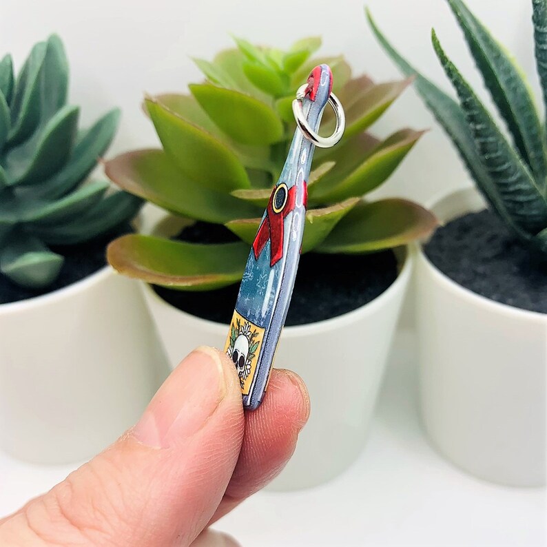 May include: A blue glass bottle charm with a skull and floral design, a red ribbon, and a silver ring at the top.
