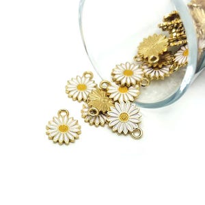 May include: Daisy flower charms in gold, white, and yellow enamel. The charms are scattered, some spilling from a clear glass container. The daisy charms have a gold outline, white petals, and a yellow center.