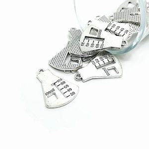 May include: A pile of silver-toned metal charms shaped like beakers with a graduated scale printed on them. The charms are 100ml beakers and have a loop at the top for attaching to a necklace or bracelet.