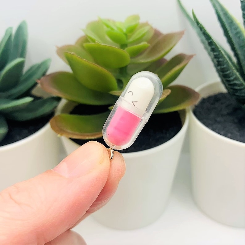 Op de afbeelding: Een transparante plastic pillenhanger met een roze en witte pil erin. De pil heeft een zwart smileygezicht erop getekend.