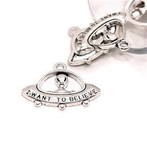 May include: Two silver charm pendants shaped like UFOs with an alien inside. The bottom pendant has the text "I WANT TO BELIEVE" on the bottom.
