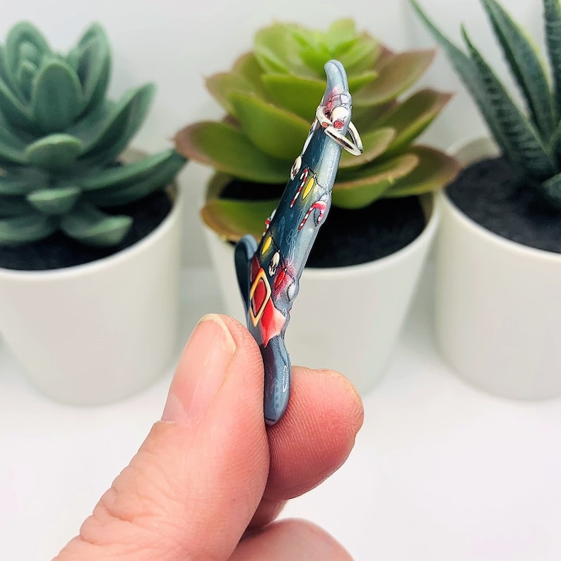 May include: A blue and red painted witch hat charm with a silver ring attached. The charm is being held by a hand.
