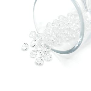 Może przedstawiać: Szklany słoik wypełniony przezroczystymi szklanymi koralikami. Koraliki wysypują się ze słoika na białe podłoże.