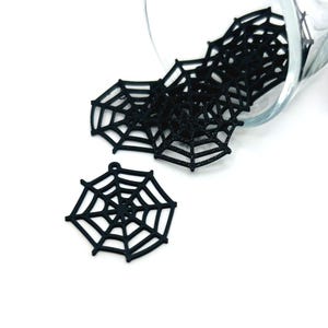Puede incluir: Encantos de telaraña negra, perfectos para manualidades de Halloween. Estas pequeñas y detalladas telarañas están hechas de un material oscuro y están dispersas sobre una superficie blanca, con algunas saliendo de un recipiente de vidrio transparente.