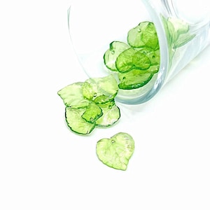 May include: A group of green glass leaf-shaped charms, some are heart-shaped, spilling out of a clear glass container.