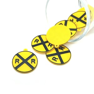 May include: Yellow and black railroad crossing charms with the letter "R" in each quadrant. The charms are round with a small loop at the top. Some charms are spilling out of a clear glass.