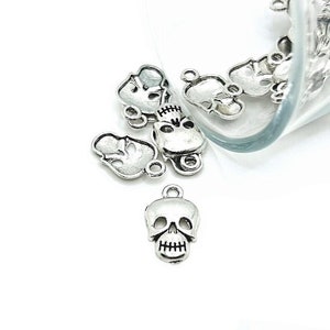 May include: A close-up of silver skull charms with a loop for attaching to jewelry or other crafts. The charms are scattered on a white surface.