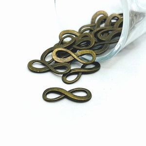 Pode incluir: Um monte de pingentes de símbolo de infinito de ouro antigo que se derramam de um frasco de vidro. Os pingentes são de metal e têm um aspeto ligeiramente envelhecido.