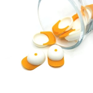May include: White and orange baseball cap-shaped candies spilling from a clear glass. The candies have a white top with a small orange dot and an orange brim. The caps are scattered on a white surface.