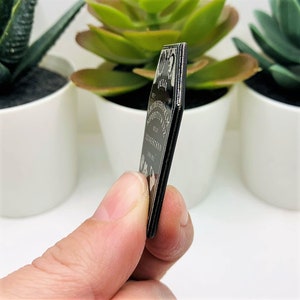 Puede incluir: Planchette de tablero Ouija de metal negro con un dise&ntilde;o grabado en plata. La planchette tiene forma de ata&uacute;d y tiene las letras del alfabeto, los n&uacute;meros y las palabras "s&iacute;", "no", "adi&oacute;s" y "hola" grabadas en ella.