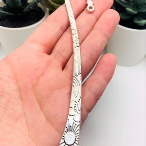May include: Silver bookmark with a sun and clouds design. The bookmark is curved and has a small loop at the top.
