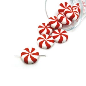 Puede incluir: Cuentas de caramelo a rayas rojas y blancas, que se asemejan a caramelos de menta, están esparcidas sobre una superficie blanca. Una cuenta está perforada con un alfiler plateado. Un vaso transparente está volcado, derramando más caramelos. Los caramelos tienen un diseño en espiral.