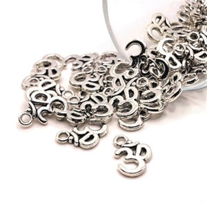 May include: A pile of silver Om symbol charms, some with a loop for hanging, spilling out of a glass container.
