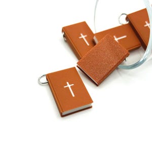 Puede incluir: Cinco pequeños amuletos en forma de libro, de color marrón, con un diseño de cruz blanca. Los amuletos están hechos de un material texturizado y tienen un anillo plateado para sujetarlos a un llavero u otros artículos.