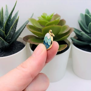 May include: Gold charm with a blue and white enamel design of waves. The charm is round and has a loop for attaching to a necklace or bracelet.