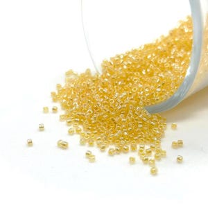 May include: A clear glass container tipped over, spilling a large quantity of small, golden-yellow seed beads onto a white surface. The beads are cylindrical and have a shiny, reflective quality.