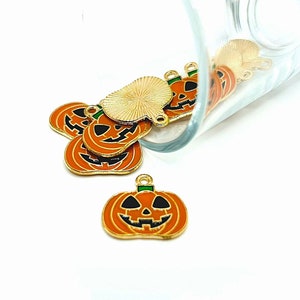 May include: A group of gold-colored metal pumpkin charms with orange enamel and black and green painted details. The charms are spilling out of a clear glass jar.