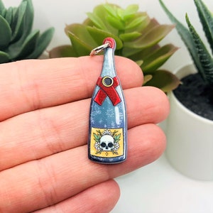 May include: A blue glass bottle charm with a skull and flowers on the label. The bottle has a red ribbon around the neck and a silver ring at the top.